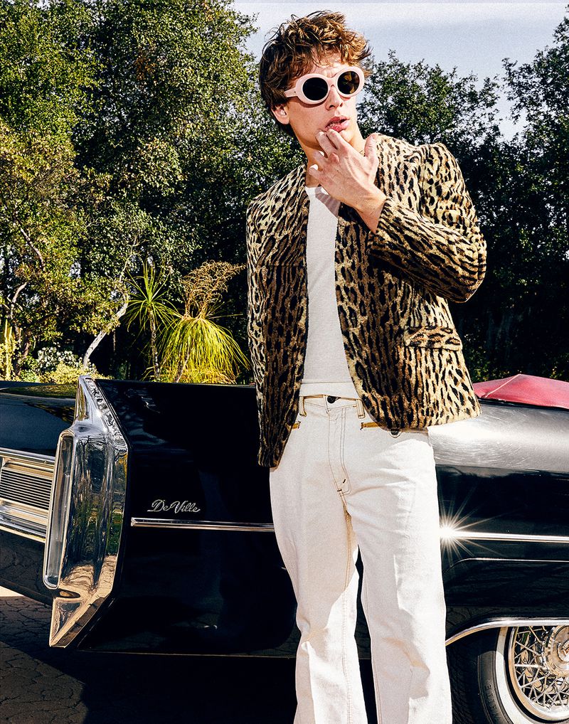 A young adult male in a leopard-print jacket poses next to a black Cadillac Deville under dappled sunlight with trees in the backdrop.