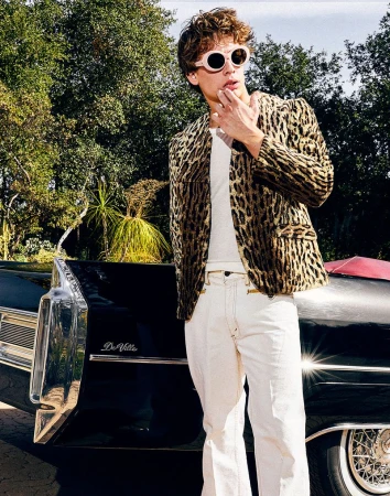 A young adult male in a leopard-print jacket poses next to a black Cadillac Deville under dappled sunlight with trees in the backdrop.