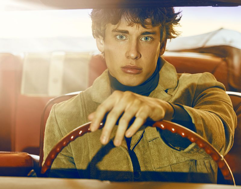 Man in car looks toward camera; he is driving a vintage car, wearing tan coat and black turtleneck, warm light, and shallow depth of field.
