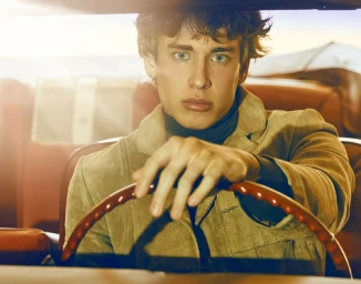 Close-up portrait of a young man holding a vintage steering wheel inside a classic car during sunset.