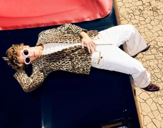 Model wearing leopard print jacket, white pants and pink sunglasses lying on top of a car against a dry cracked earth background.