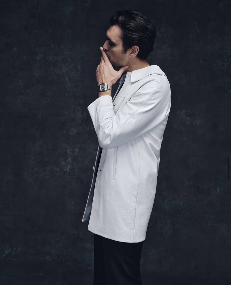 Profile view of a man in a white coat with dark hair, hand near his face, wearing a watch against a dark textured backdrop.