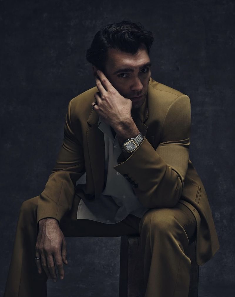 Portrait of a handsome adult male in a tan suit, seated and gazing at the camera with hand near his face, set against a dark backdrop.