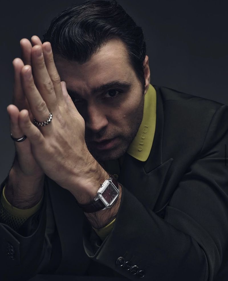 Portrait of a dark-haired adult male wearing a suit with a bright yellow collar with clasped hands in a dimly lit photo studio.