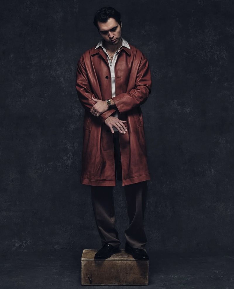 Full-length studio shot of a man in a brown leather coat and trousers posing on a box against a mottled grey backdrop.