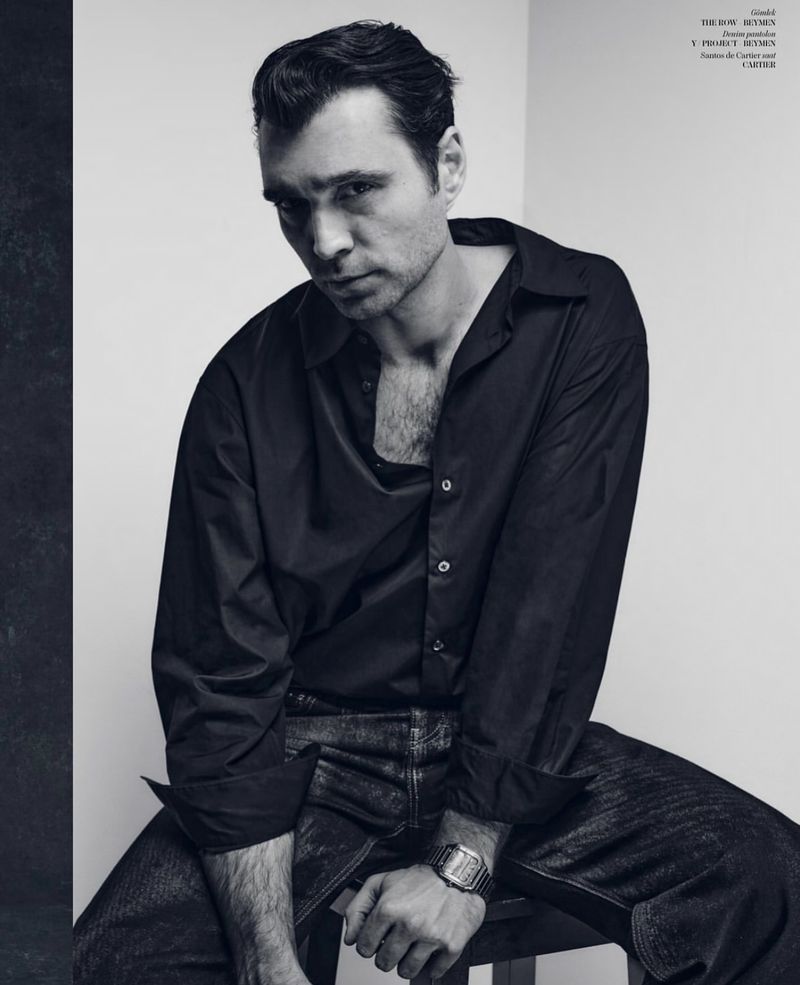 Portrait of a male model in a black shirt and jeans sitting on a stool in a photo studio with soft lighting.
