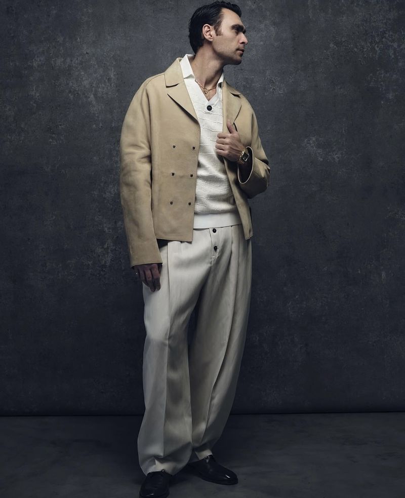 Hakan Kurtaş posing in a beige double-breasted jacket and wide-leg cream trousers against a dark textured backdrop.