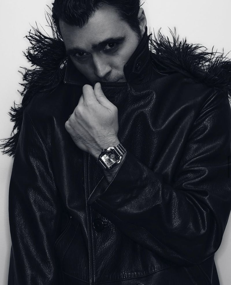 Monochrome portrait of a man in a leather jacket with feathered shoulders, looking at the camera with a serious expression indoors.