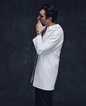 Man wearing a white coat and silver wristwatch posing in profile against a textured dark grey background