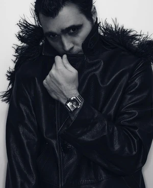 Hakan Kurtas wearing a black leather coat with feather collar and pulling up the lapel against a light studio backdrop.