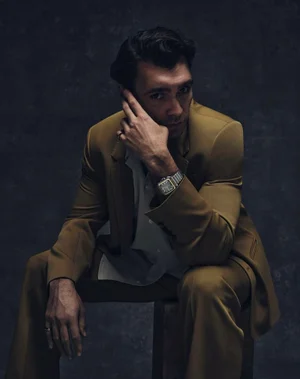 Hakan Kurtas seated wearing an ochre suit, open shirt, and silver watch against a dark textured backdrop.
