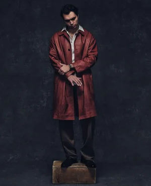 Man wearing a long red leather coat and collared shirt standing on a wooden box against a dark textured background