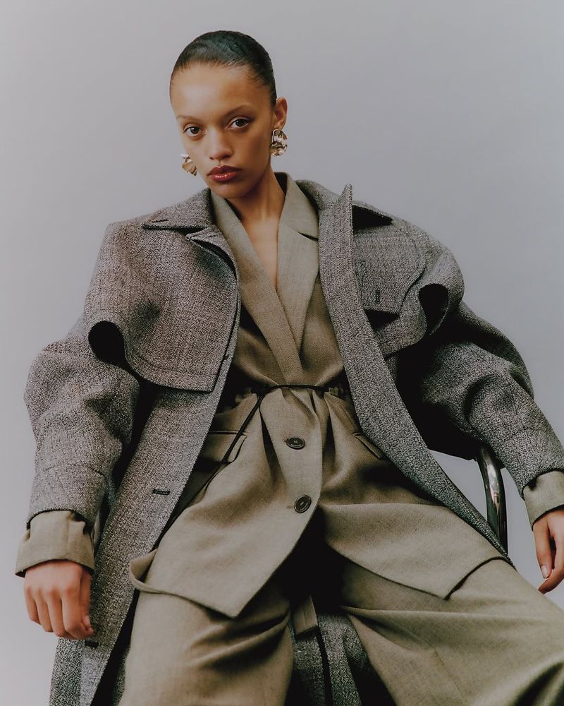 Fashion editorial shot of a woman in a tweed overcoat and tan suit, posing in a studio setting. Medium shot, eye-level angle.