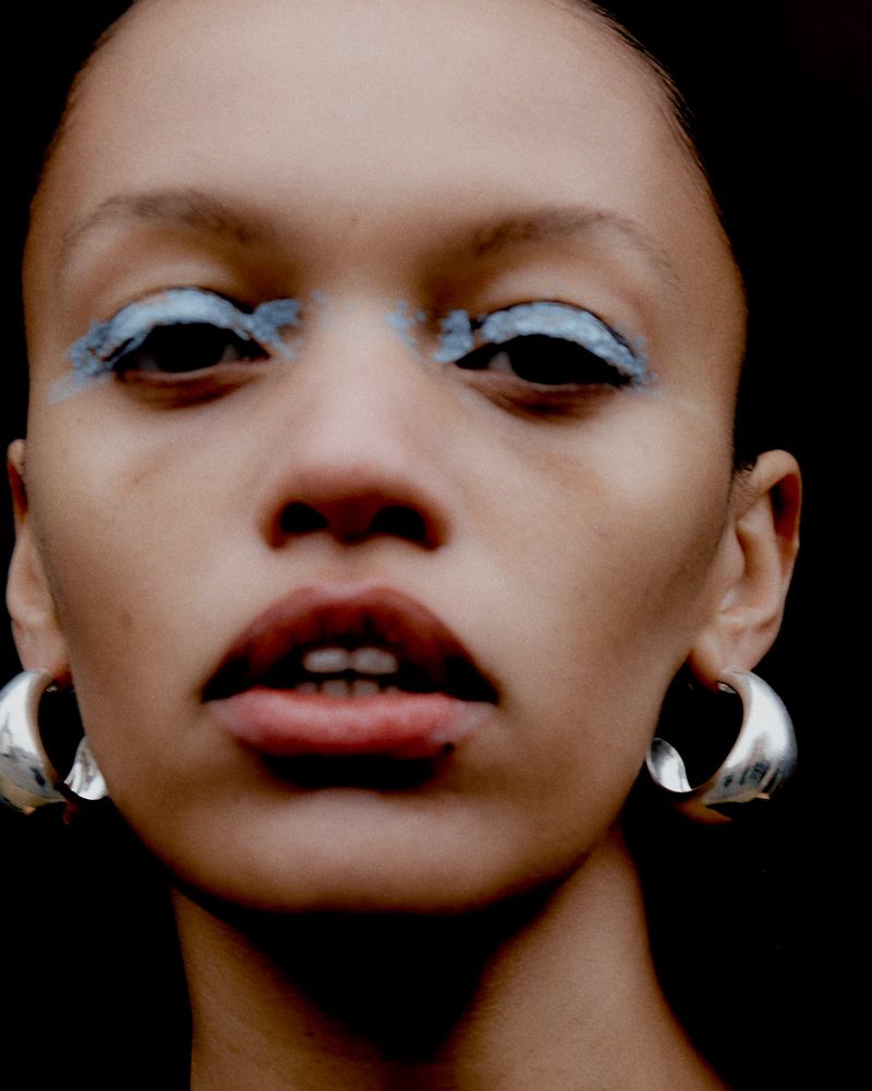 Close-up portrait of a woman with light skin, bright blue eyeshadow, and hoop earrings against a dark, contrasting backdrop.