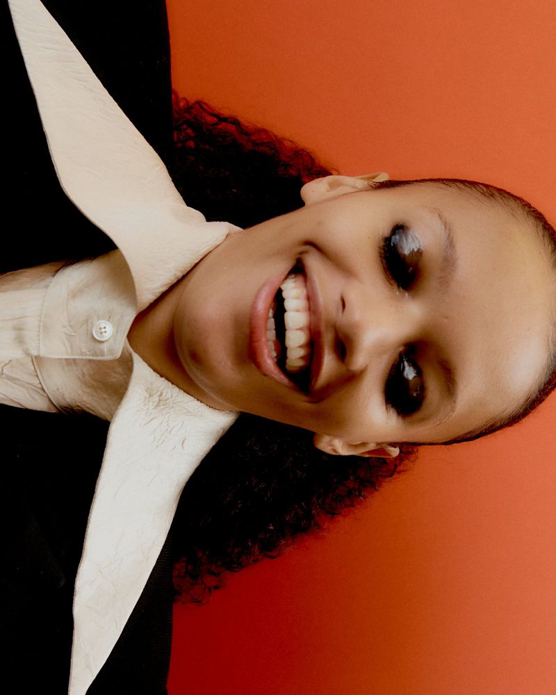 Close-up portrait of Kukua Williams smiling in a black jacket and pointed cream collar against an orange studio backdrop.
