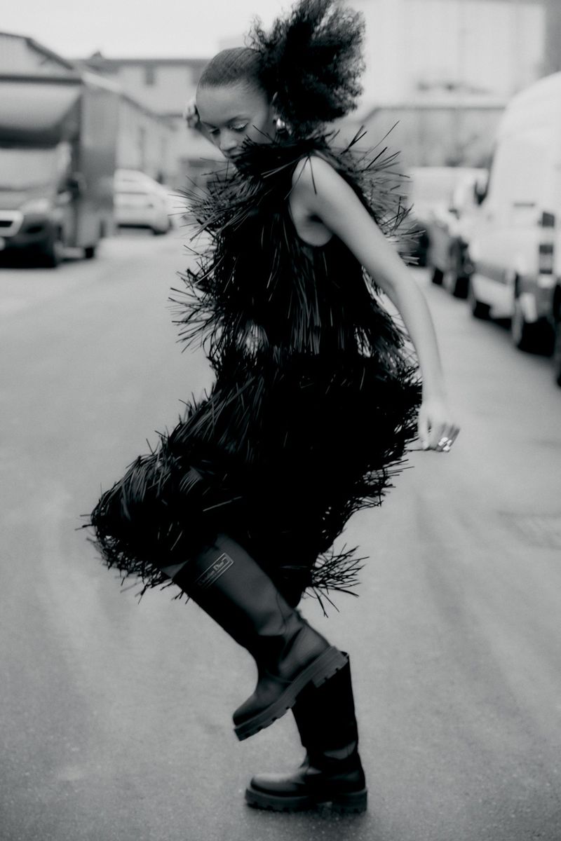Fashion editorial shot of an African female model in a black fringe dress and boots posing on a city street with parked vans.