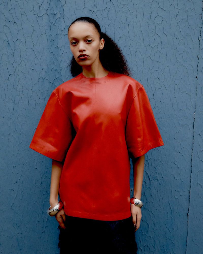 A young adult female poses against a textured blue wall wearing a red, short-sleeved top and bracelets for a fashion shot.