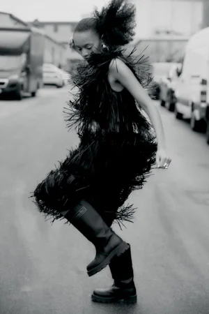 Model Kukua Williams wearing a black feathered fringe dress and chunky black boots dancing on an urban street in black and white.