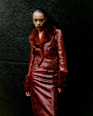 Model Kukua Williams wearing a burgundy snakeskin jacket and matching skirt with fur trim against a dark textured wall.