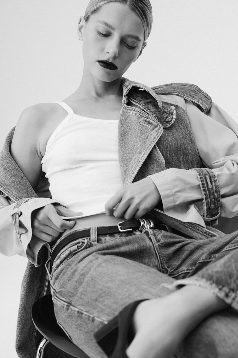 Black and white shot of a fair-skinned woman in a tank top, jeans and a denim jacket on her shoulder sitting in a photo studio.