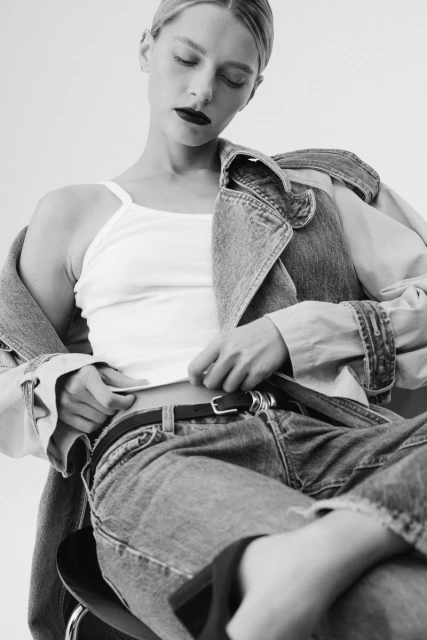 Black and white shot of a fair-skinned woman in a tank top, jeans and a denim jacket on her shoulder sitting in a photo studio.
