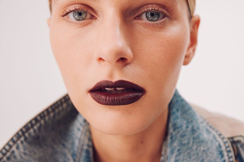 Close-up beauty portrait of model Gray Corton wearing dark burgundy lipstick and an acid-wash denim jacket.
