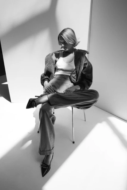 Monochrome fashion shot of a woman in a jacket, tank top, and pants, sitting on a chair in a studio with strong lighting and shadows.
