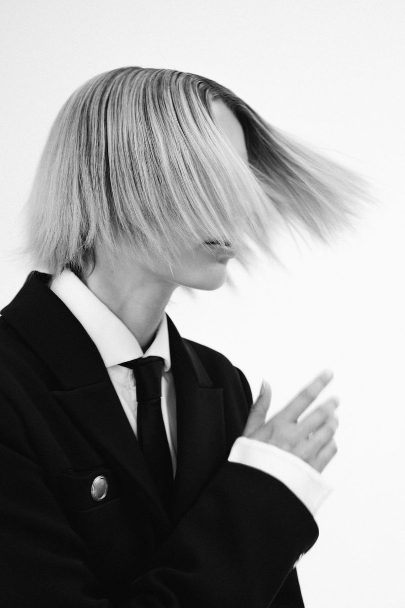 Monochrome portrait of a person with flowing blond hair, wearing a black blazer, white collared shirt, and black tie against a white backdrop.
