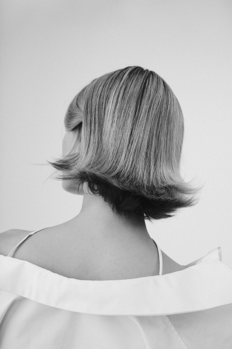 Black and white back view portrait of a person with a retro flipped short bob haircut wearing an off the shoulder white garment.