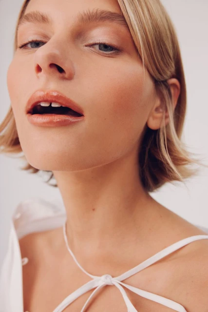 Close-up portrait of a blonde woman with glossy lips and a white top, showcasing beauty and fashion in a studio setting.
