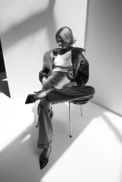 Model seated on a metal chair wearing a shearling-lined jacket, white tank top, and trousers in a studio setting