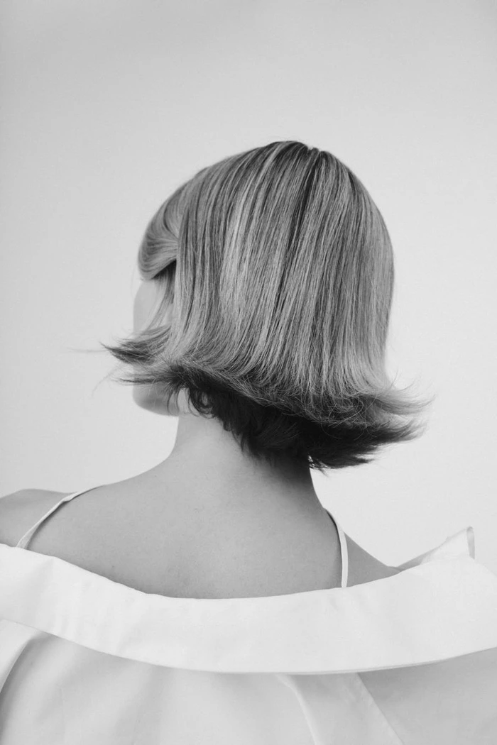 Black and white back view portrait of a person with a retro flipped short bob haircut wearing an off the shoulder white garment.