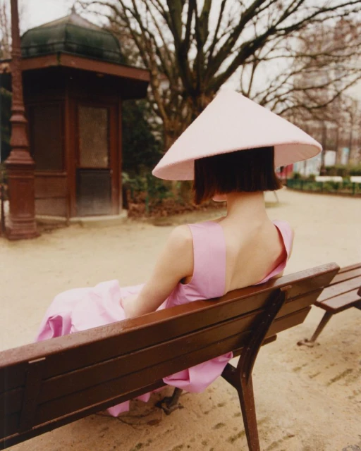 Woman with bob haircut sits on park bench wearing pink dress and wide-brimmed conical hat with park shelter and bare trees behind.