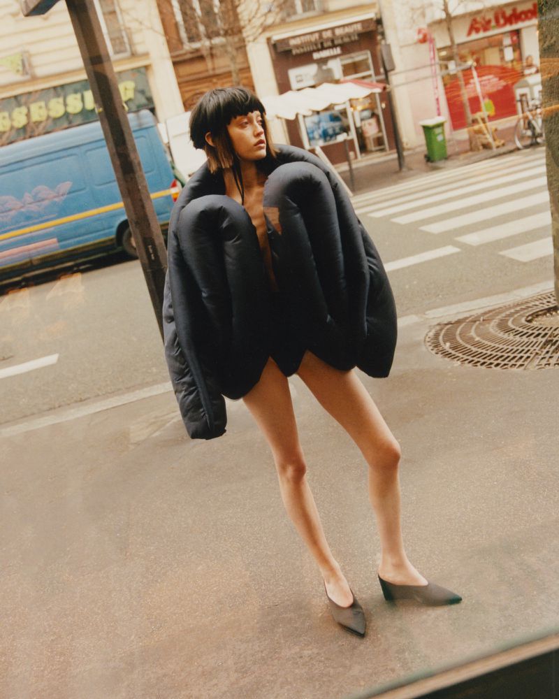 An adult female model in a black puffer coat and shoes poses on a city street with buildings and a blue van in the background.