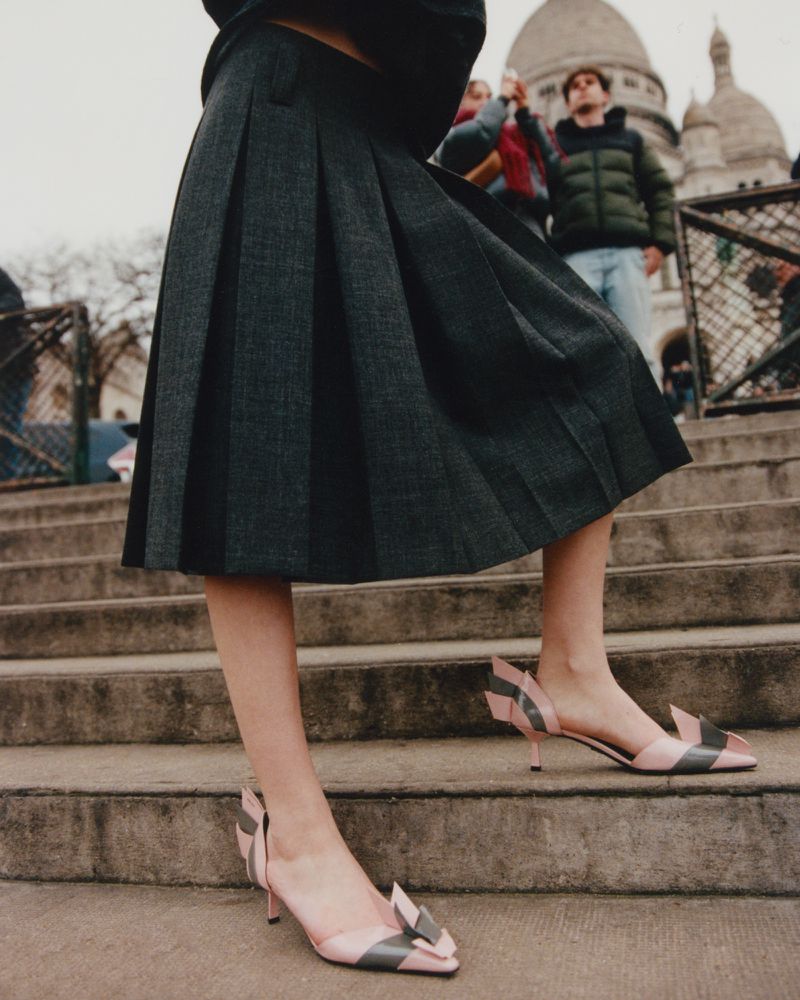Woman in pleated skirt and pink heels walks on stairs in front of Sacré-Cœur Basilica in Paris, fashion editorial shot.