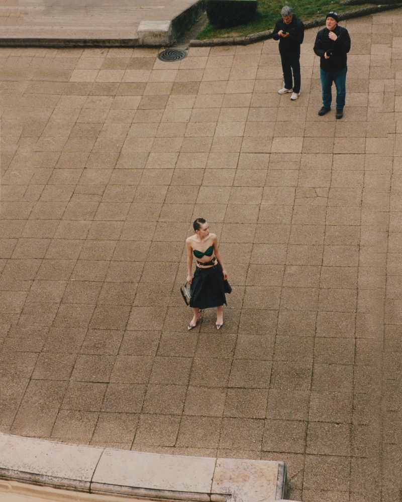 High-angle shot of a stylish woman in a bra top and skirt on a tiled surface, observed by two men in the background outdoors.