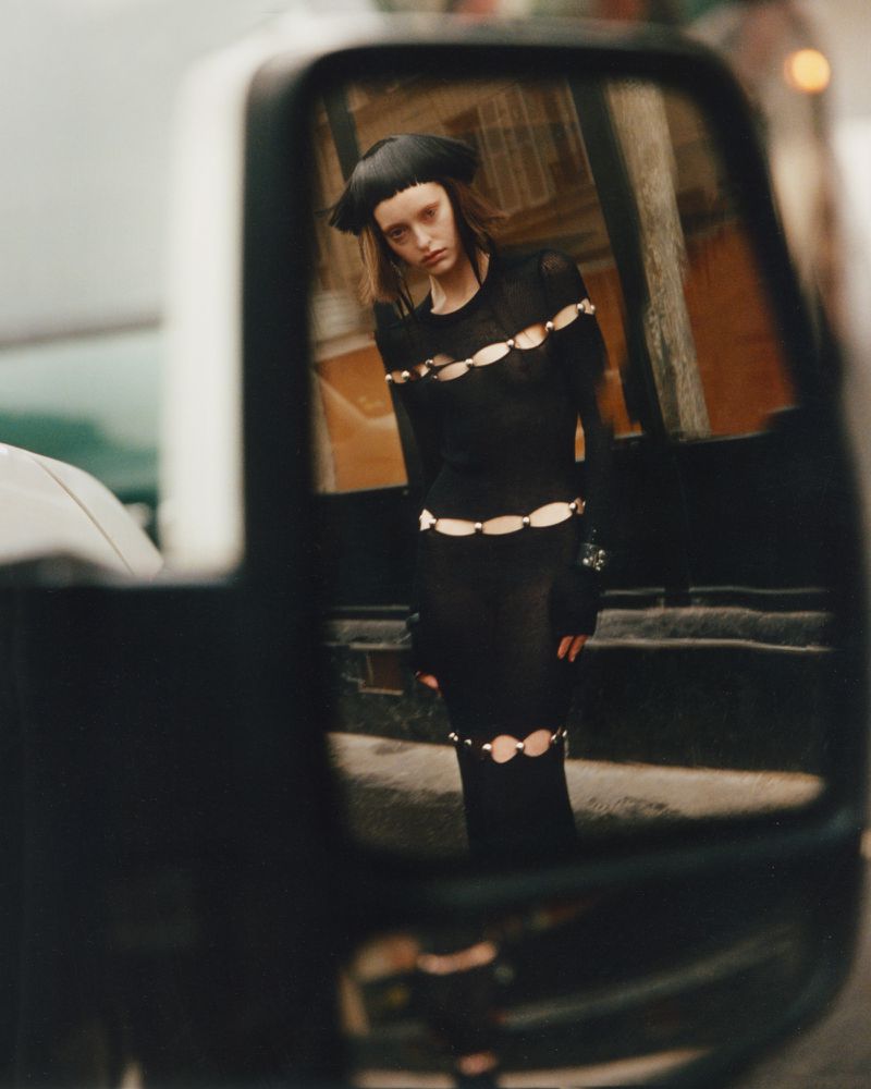 A fashionable young woman reflects in a side-view car mirror, wearing a black dress with cut-out details and a matching black hat.