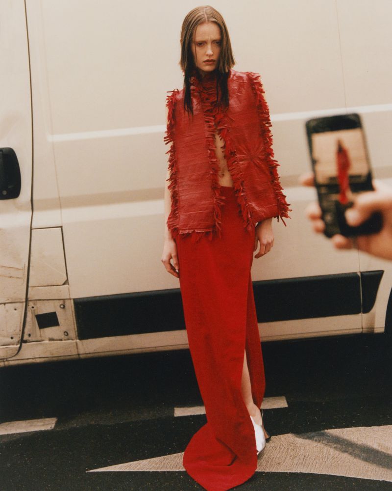A woman in a red dress and matching vest stands in an urban environment next to a white van while someone takes a photo with a cell phone.