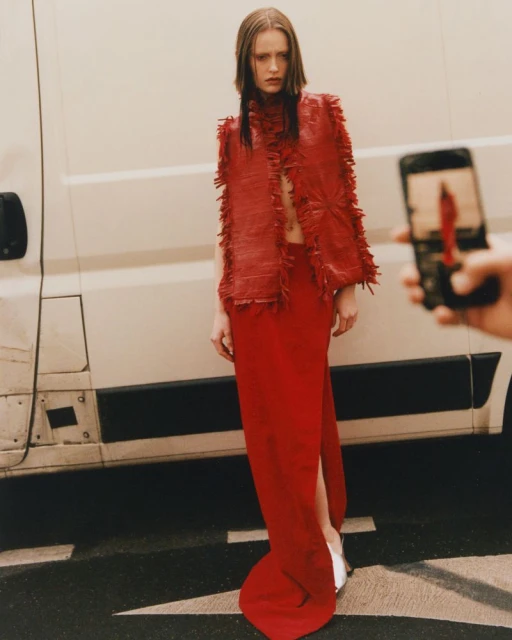 A woman in a red dress and matching vest stands in an urban environment next to a white van while someone takes a photo with a cell phone.