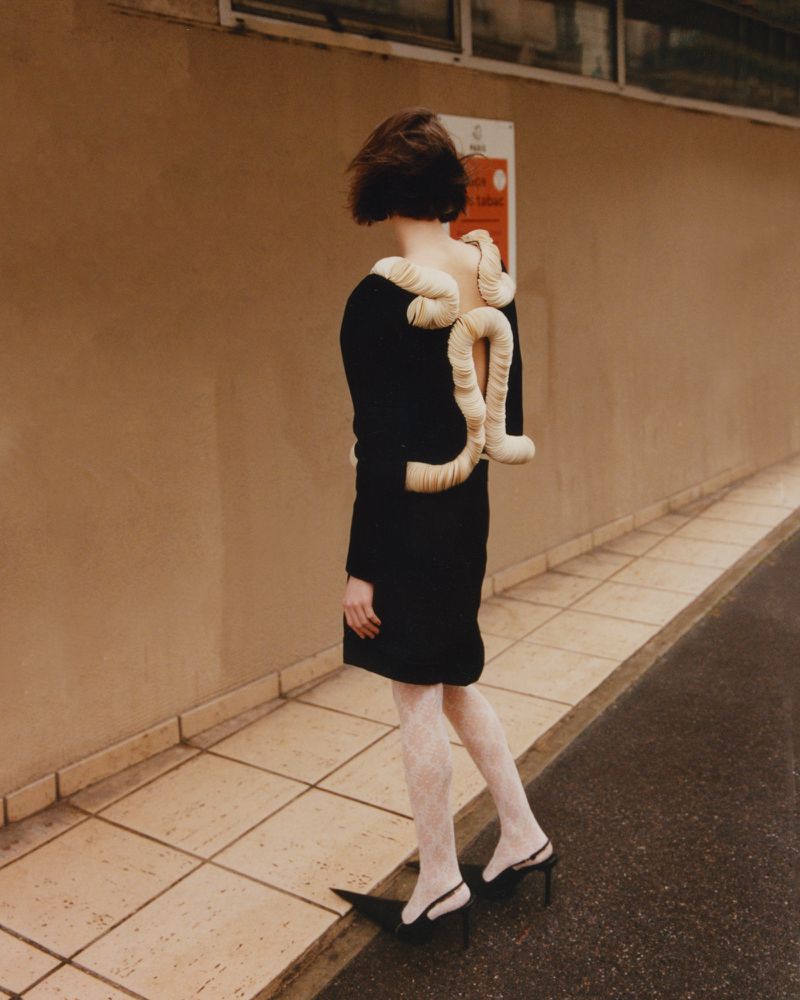 Model Avah Adams stands facing away wearing a black open-back dress with sculptural cream accents and white patterned tights outdoors.