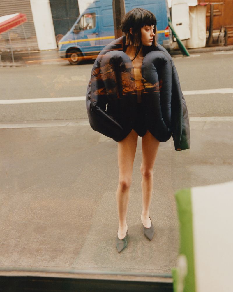 A woman with dark hair stands on an outdoor city street wearing an oversized, padded jacket, visible legs, and dark shoes.