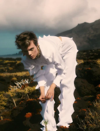A young adult bends forward in a white, frilled outfit on a lava field, beneath mountains and a cloudy sky for a fashion editorial.