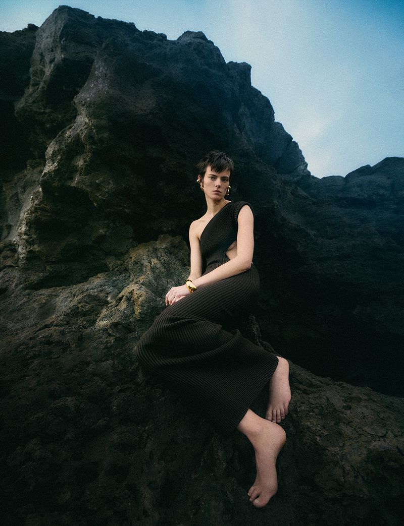 A woman in a black dress poses on dark rocks with a cloudy sky background, showcasing fashion and editorial style.