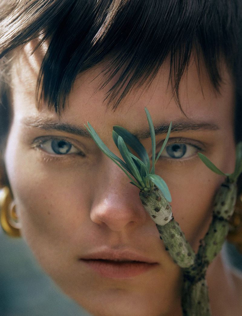 Close-up portrait of a young woman with short brown hair, fair skin, and blue eyes, holding a plant stem with green leaves near her face.