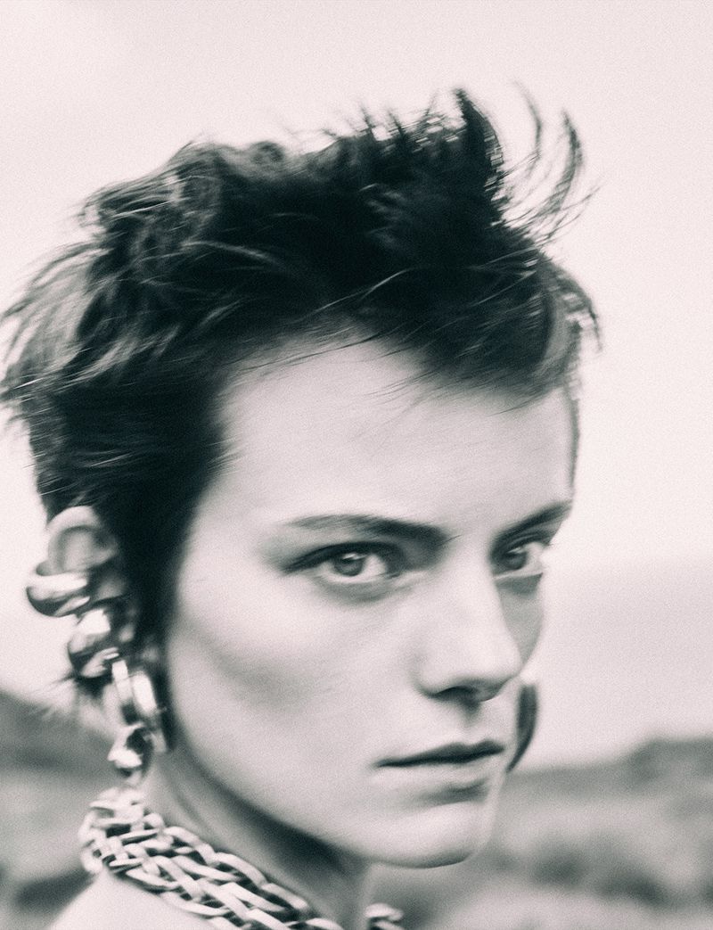 Black-and-white portrait of an androgynous adult with short hair, statement earrings, and a thick chain necklace, set outdoors.