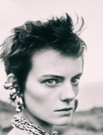 Black-and-white portrait of an androgynous adult with short hair, statement earrings, and a thick chain necklace, set outdoors.