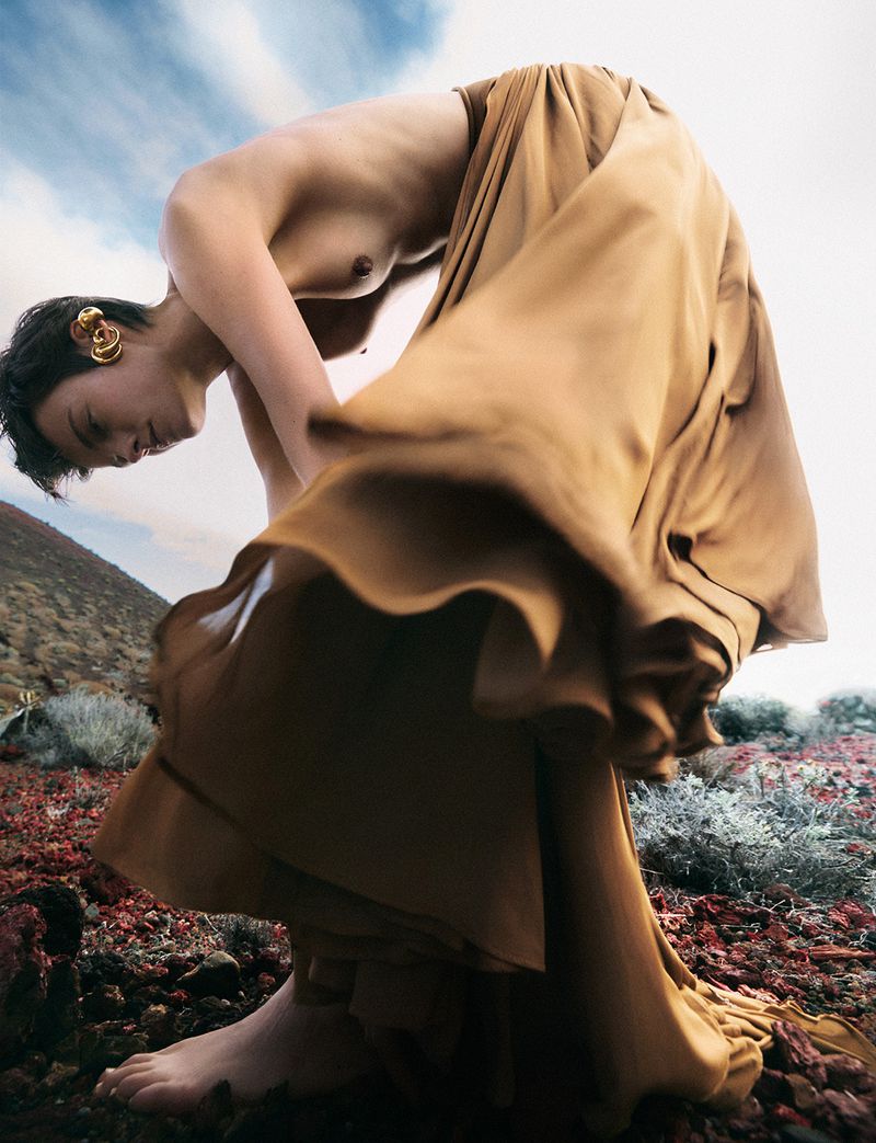 Female fashion model posing in gold dress on a mountain slope. Barefoot woman with short dark hair wears golden ear cuff. Sky background.