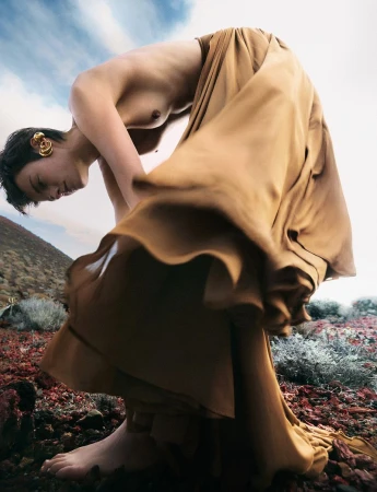 Female fashion model posing in gold dress on a mountain slope. Barefoot woman with short dark hair wears golden ear cuff. Sky background.