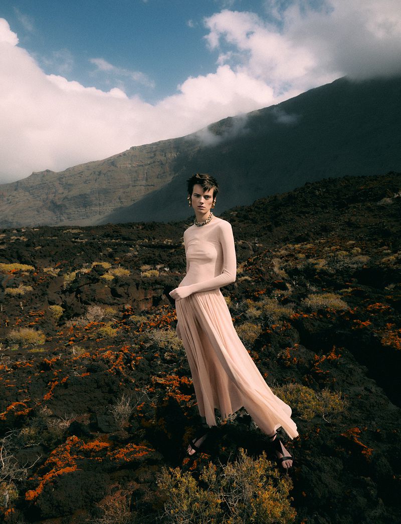 A woman in a long dress poses in a stark landscape with a mountain backdrop under a blue, partly cloudy sky in an outdoor fashion shot.