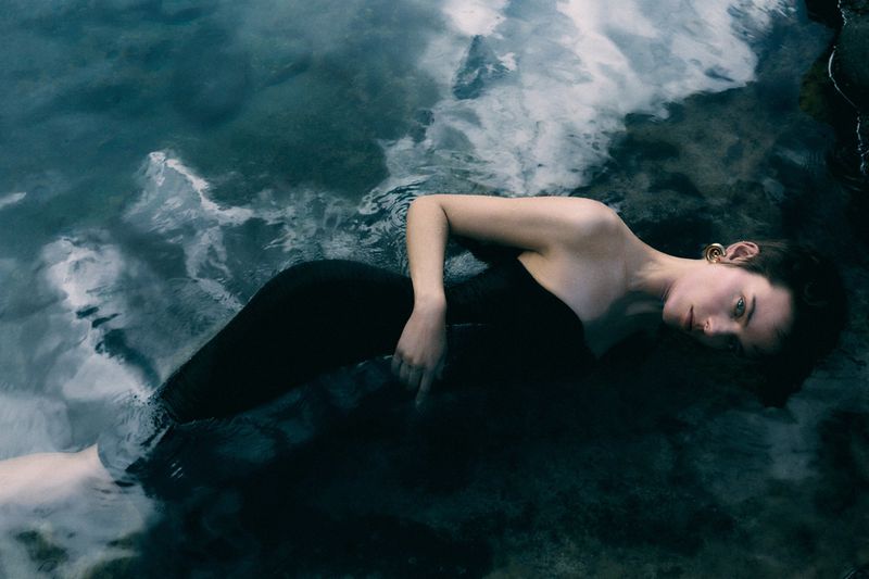 An adult female lies in water wearing a dark dress, reflecting the sky, with a shallow depth of field and low angle. Soft lighting.